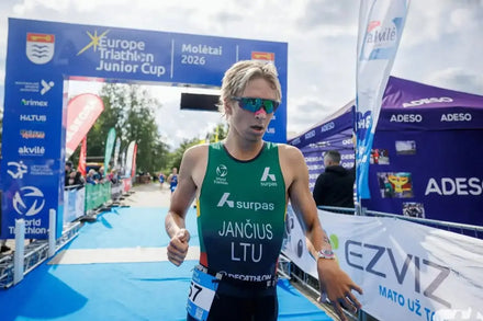 A male athlete wearing a green and black surpas athletic vest and blue sunglasses crosses a blue finish line at the Europe Triathlon Junior Cup in Molátal 2026. The background features event banners, tents, and spectators.